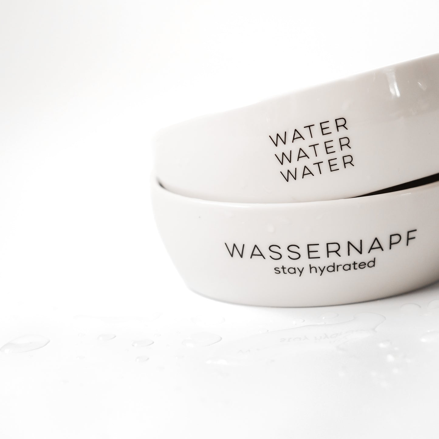 Zwei weiße Keramiknäpfe gestapelt vor einem weißen Hintergrund. Die Wassernäpfe für Katzen haben die Aufdrucke "water water water" und "Wassernapf - stay hydrated".