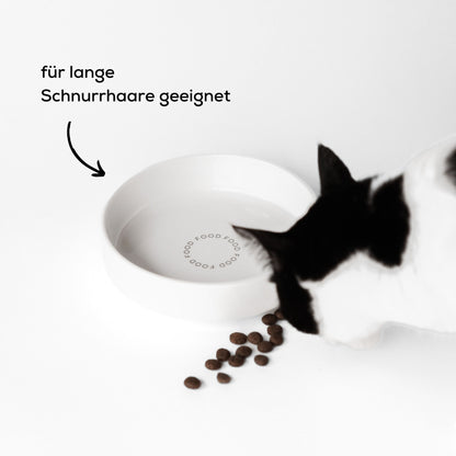 Eine schwarz weiße Katze frisst aus einem großen Futternapf aus Keramik. Vor dem Napf liegt Trockenfutter. Der Napf ist für lange Schnurrhaare geeignet.
