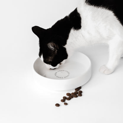 Eine schwarz weiße Katze frisst aus einem großen Futternapf aus Keramik. Vor dem Napf liegt Trockenfutter.