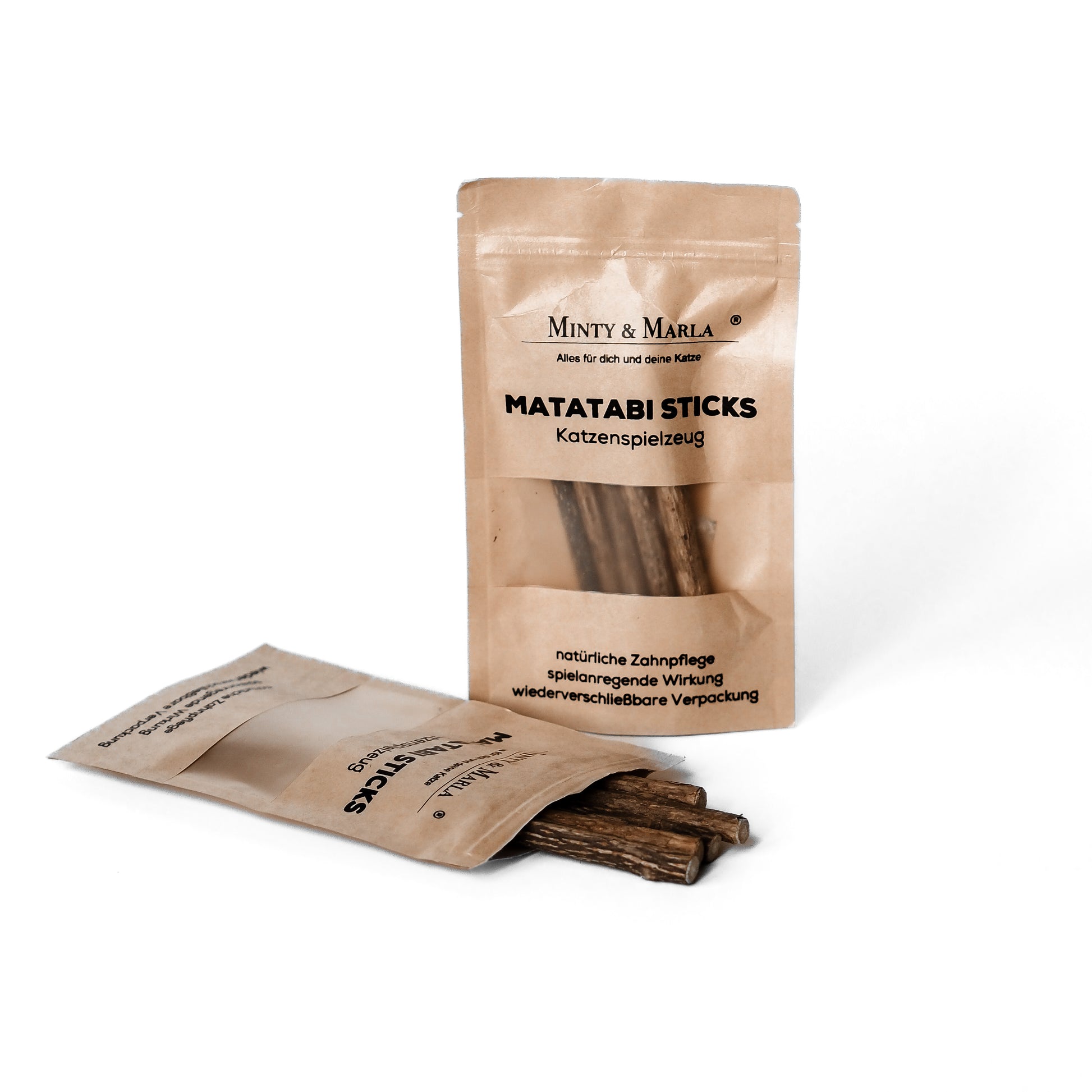 Eine braune Papierverpackung mit dem Aufdruck "Matatabi Sticks Katzenspielzeug" der Marke Minty & Marla  steht vor einem weißen Hintergund. Vor der Verpackung liegen kleine Hölzer, welche Matatabi Sticks heißen und als Spielzeug für Katzen dienen.