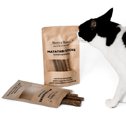 Eine braune Papierverpackung mit dem Aufdruck "Matatabi Sticks Katzenspielzeug" der Marke Minty & Marla  steht vor einem weißen Hintergund. Vor der Verpackung liegen kleine Hölzer, welche Matatabi Sticks heißen und als Spielzeug für Katzen dienen. Eine Katze riecht an den Matatabi Sticks.