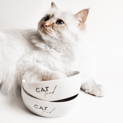 Eine helle Katze liegt hinter zwei ineinander gestapelten weißen Keramiknäpfen für Katzen mit dem Aufdruck "Cat Food".