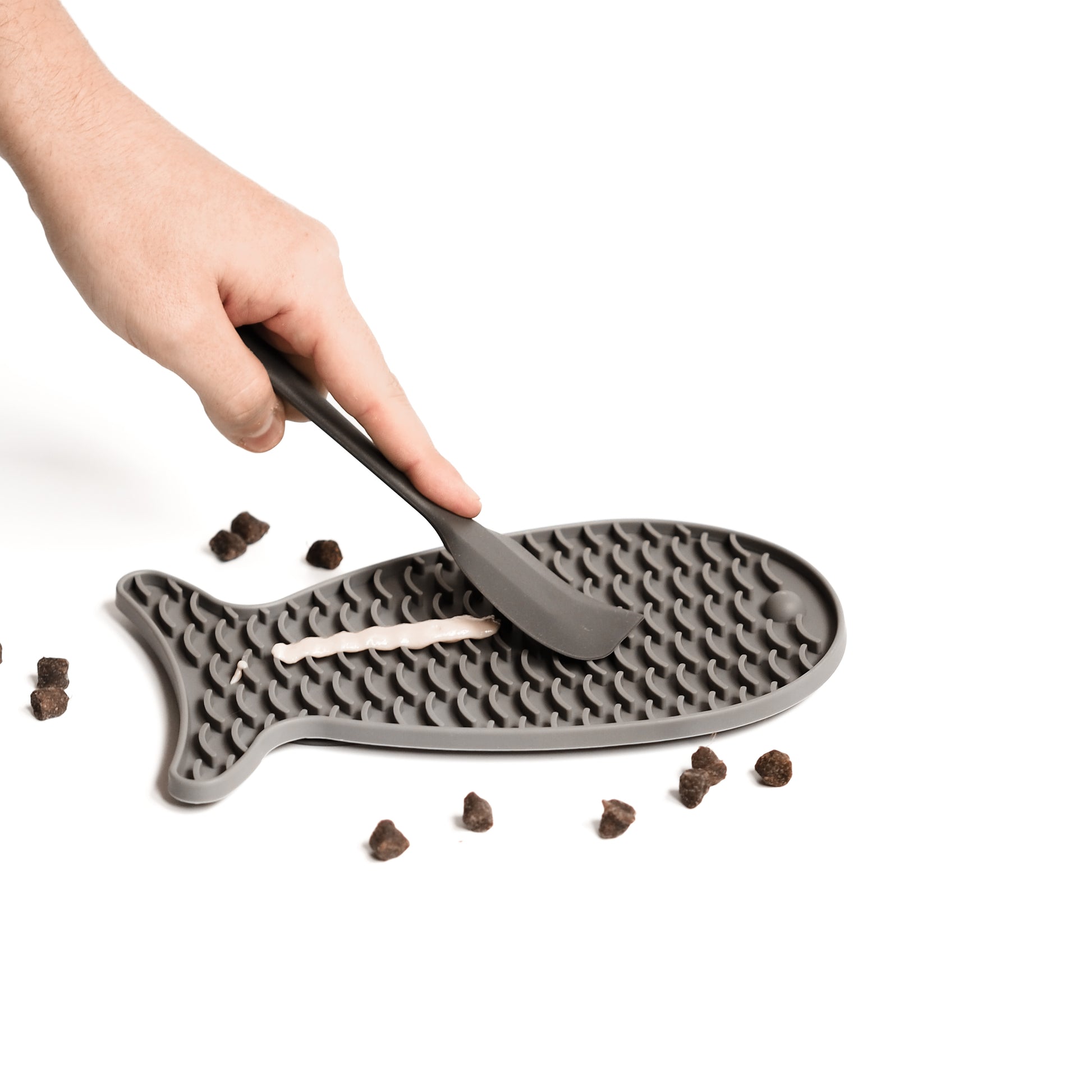 Eine menschliche Hand streicht eine Paste für Katzen auf eine graue Schleckmatte für Katzen. Neben der grauen Schleckmatte liegen Leckerlis für Katzen.