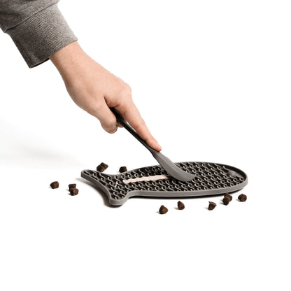 Eine männliche hand bestreicht eine graue Schleckmatte für Katzen mit Katzenfutter. Neben der Schleckmatte liegt Trockenfutter für Katzen.